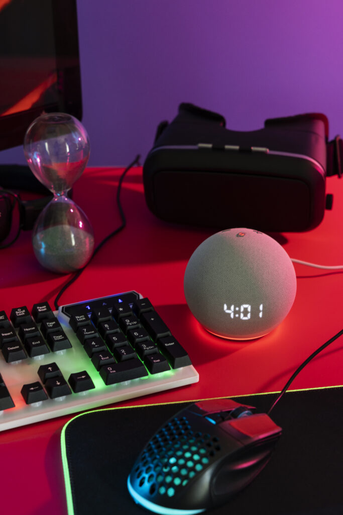 Configuración de escritorio con teclado RGB, ratón ergonómico, reloj inteligente con pantalla LED, gafas de realidad virtual y reloj de arena. Setup ideal para gamers y entusiastas tecnológicos